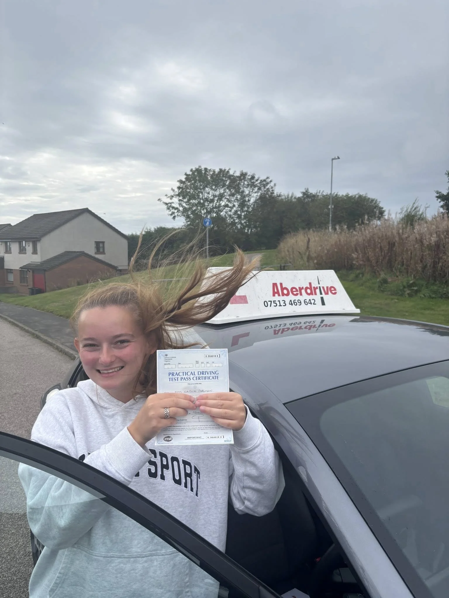 A young woman holding a practical driving test pass certificate while smiling, standing outside a car with a driving school sign on the roof, on a cloudy day.