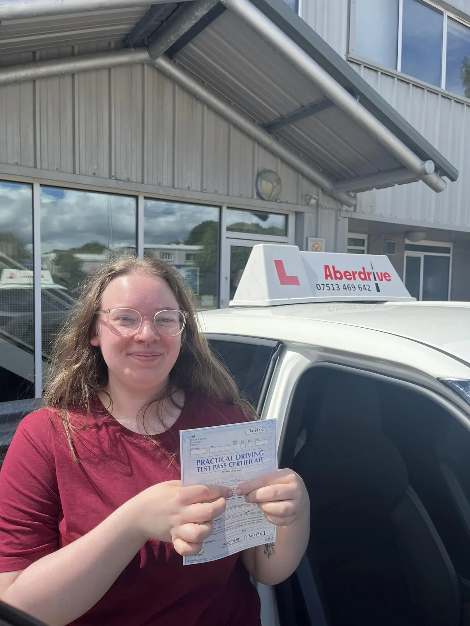 A woman with glasses and a maroon shirt holding a practical driving test pass certificate, standing beside a white car with an Aberdrive driving school sign on top, outside a building with large windows.