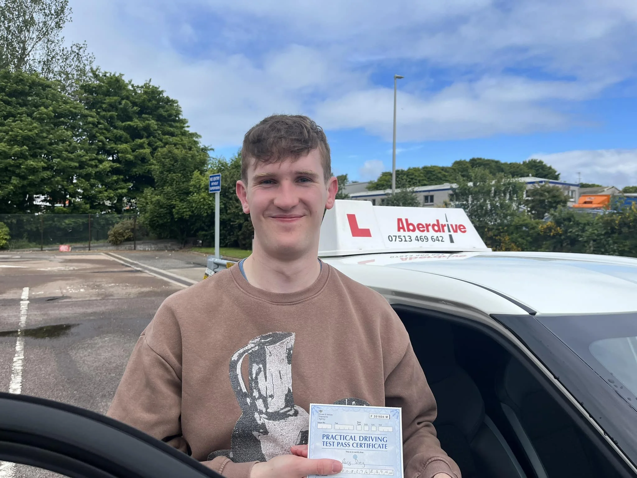 Young man holding a practical driving test pass certificate stands next to a white driving school car with a sign that reads "Aberdrive" in a parking lot with trees and a building in the background.