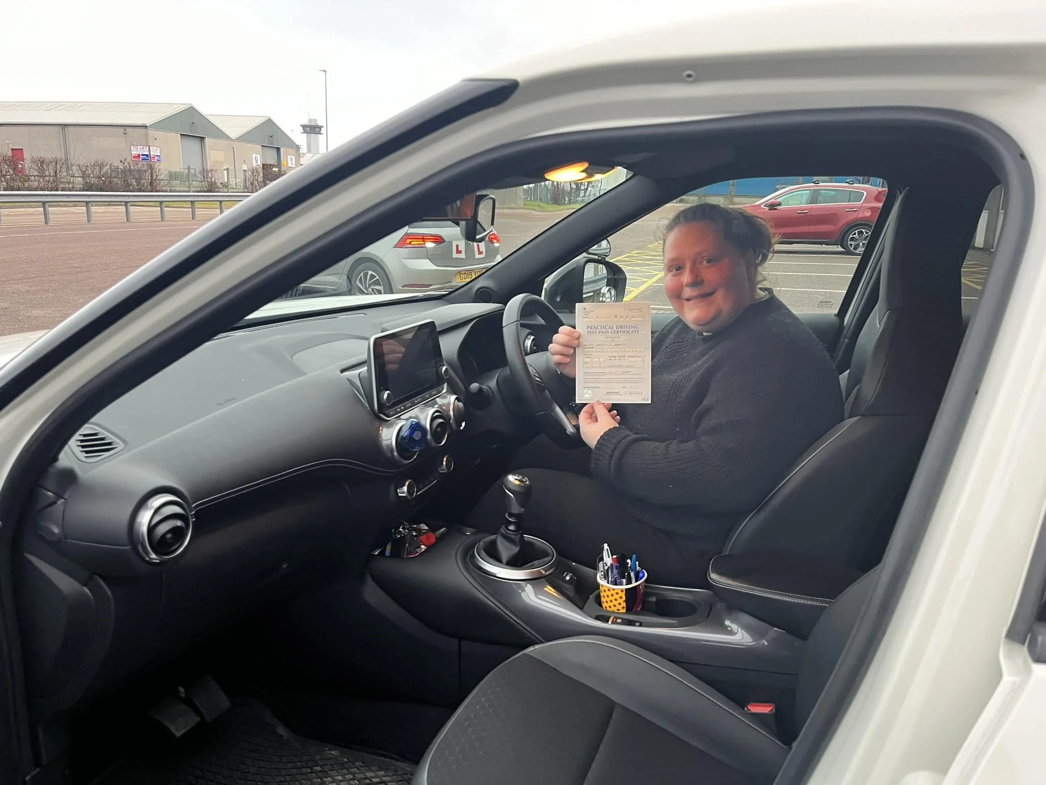 A smiling woman sitting in the driver's seat of a car, holding a practical driving test pass certificate, inside a parking lot with other vehicles and a building in the background.