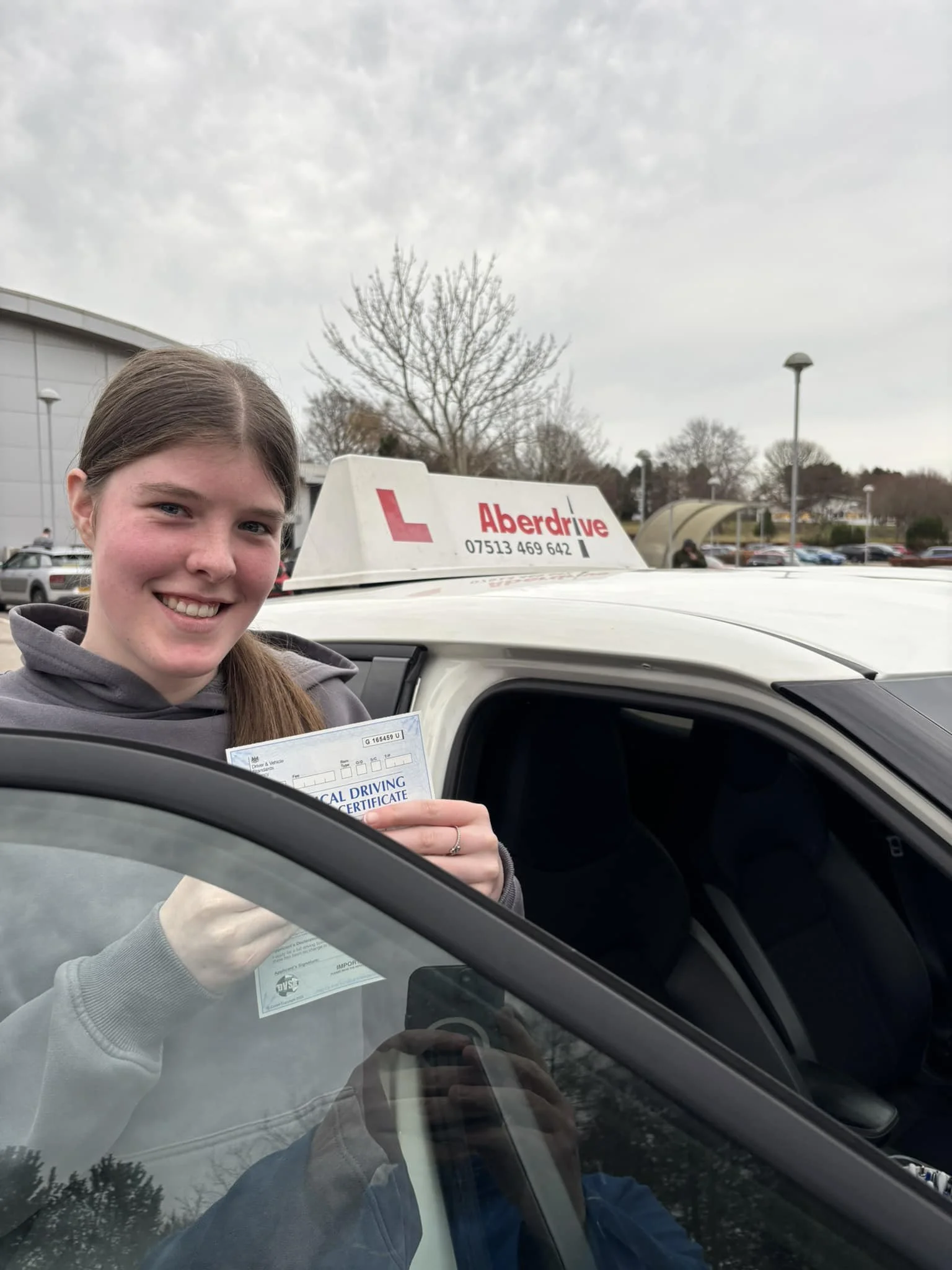 A young woman smiling and holding a newly earned driver's license in front of her while sitting in a car, with a driving school car visible behind her.