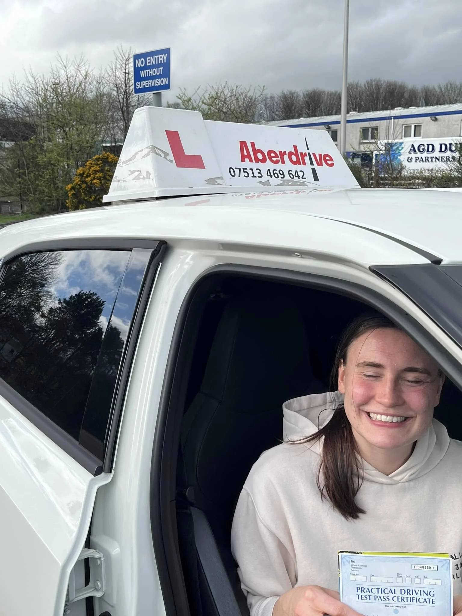 A smiling young woman sitting in the driver seat of a white driving school car, holding a practical driving test pass certificate. The car has a sign on the roof that reads 'Aberdrive' along with a phone number. The background shows trees, a sign tha
