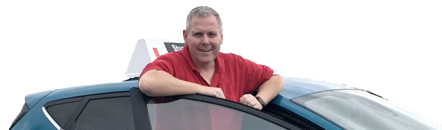 A man in a red shirt leaning out of the driver's side window of a car, smiling, with a sign in the background.