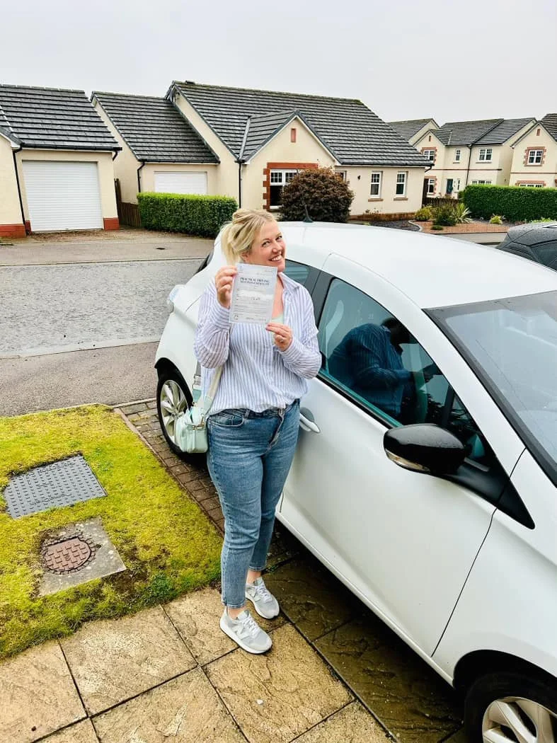 Woman smiling and holding a document next to a white car parked in a suburban neighborhood.