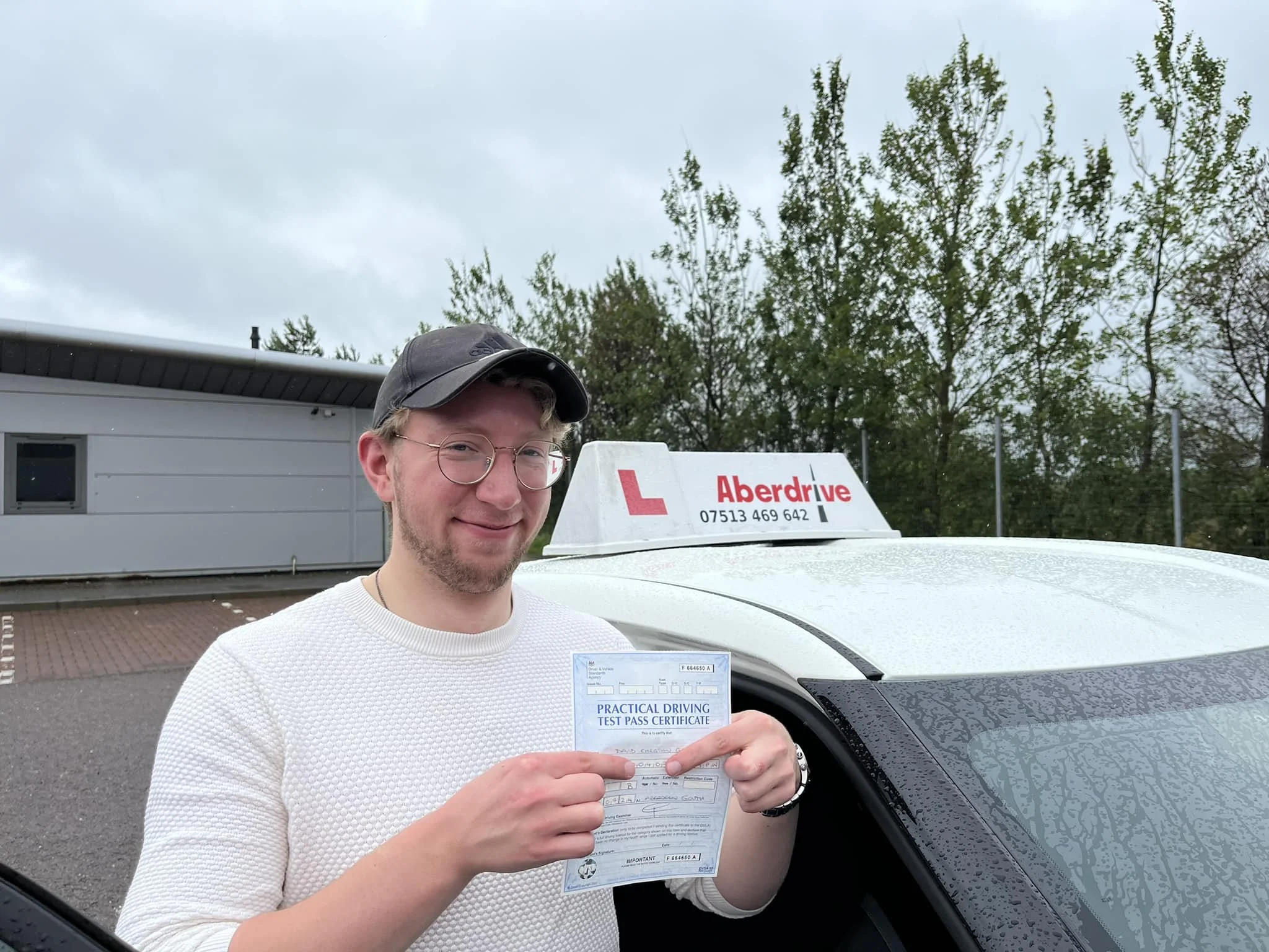 A young man with glasses, wearing a black cap and white sweater, holding a driving test pass certificate, standing next to a white driving school car with 'Aberdrive' sign on top, outdoors with trees and cloudy sky in the background.