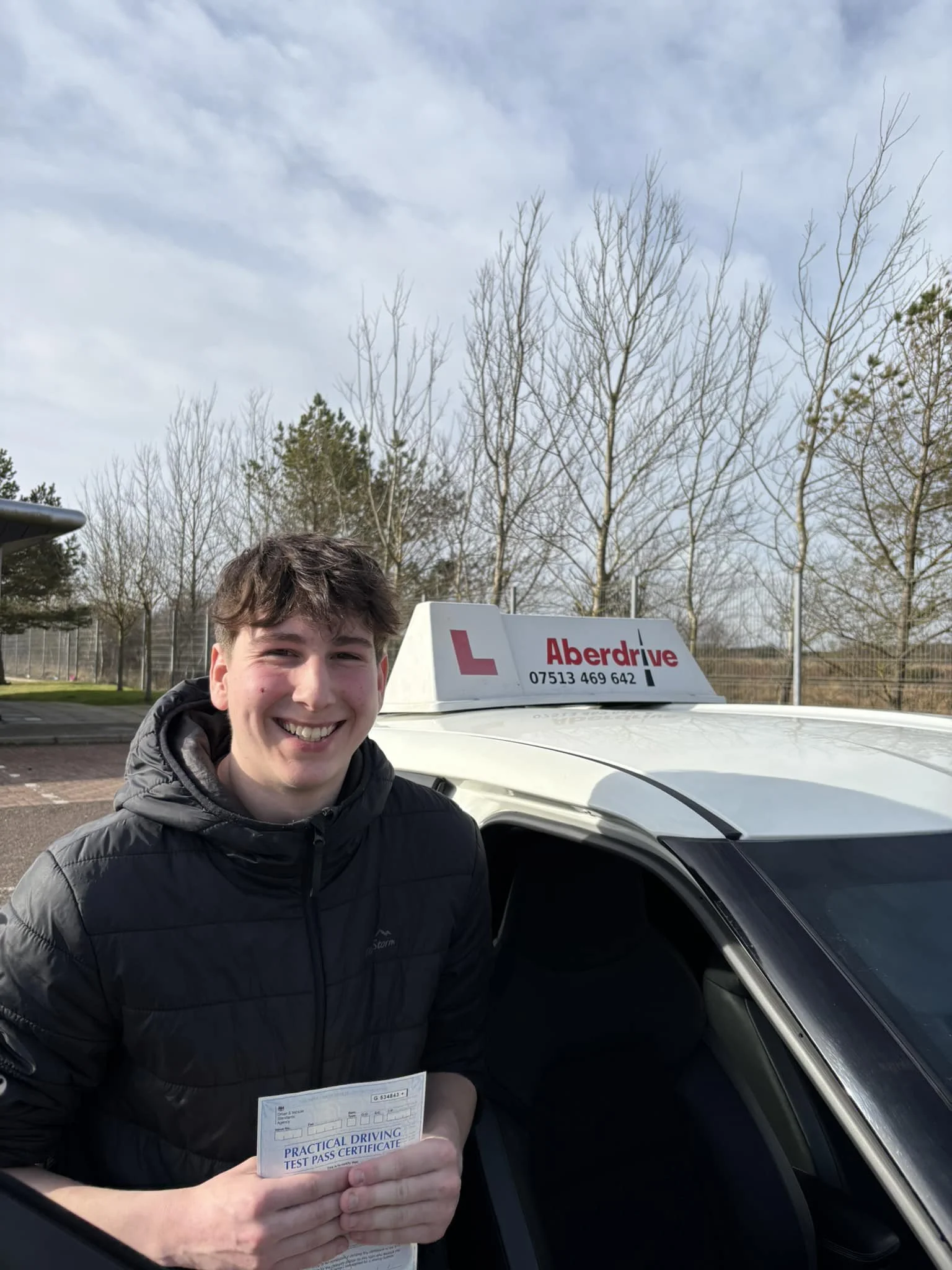 A young man smiling and holding a practical driving test pass certificate standing next to a white car with a sign on top that reads 'Aberdrive' and includes a phone number. The background shows trees and a partly cloudy sky.