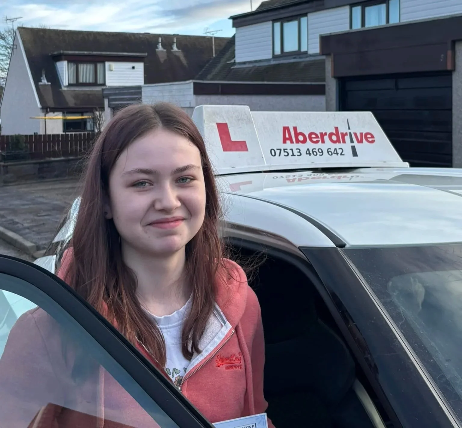 A young woman with long brown hair and a light smile is standing outside a car, which is an driving instructor's vehicle. The car has a sign on top that reads 'Aberdrive' with a red 'L' and a phone number, indicating it is used for driving lessons. Residential houses are visible in the background.
