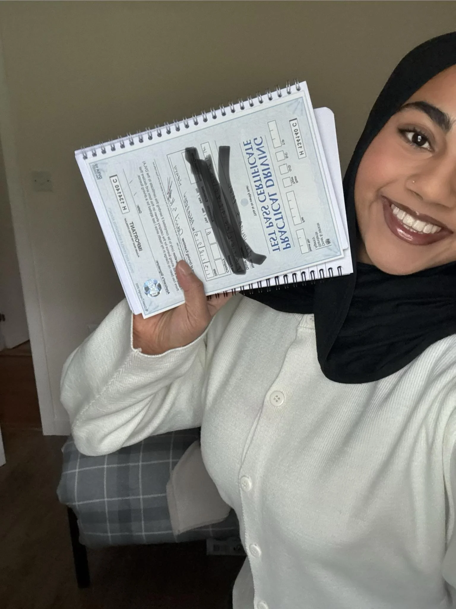 A smiling woman wearing a black headscarf and white cardigan holding a notebook with writing and black markers, showing a license or document.