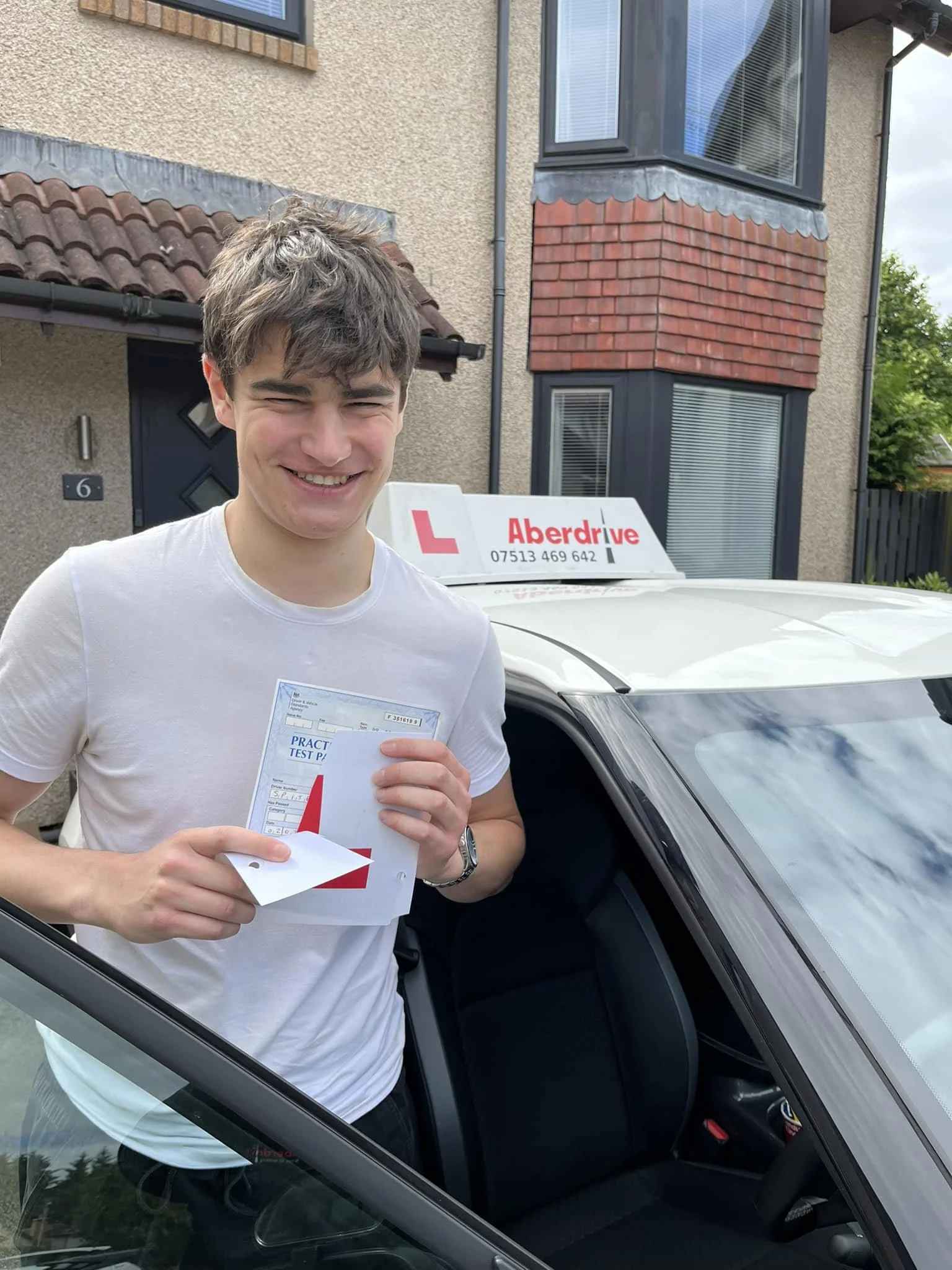 A young man smiling and holding a practical test paper and documents in front of a car with a driving school sign on the roof, outside a residential house.
