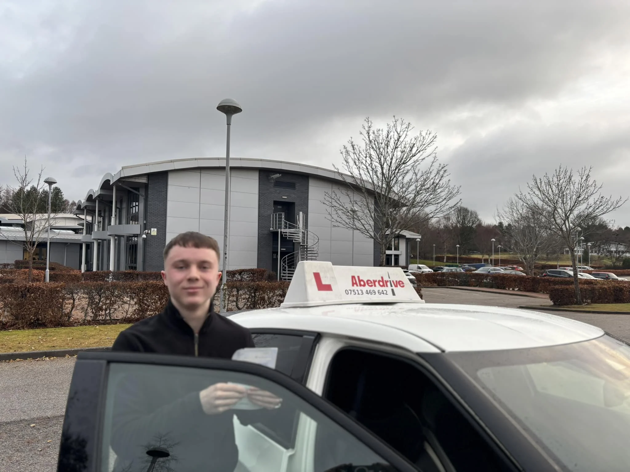 A young man standing next to a driving school car with a sign on top that reads 'Aberdrive' in a parking lot with leafless trees and a large, modern building in the background.