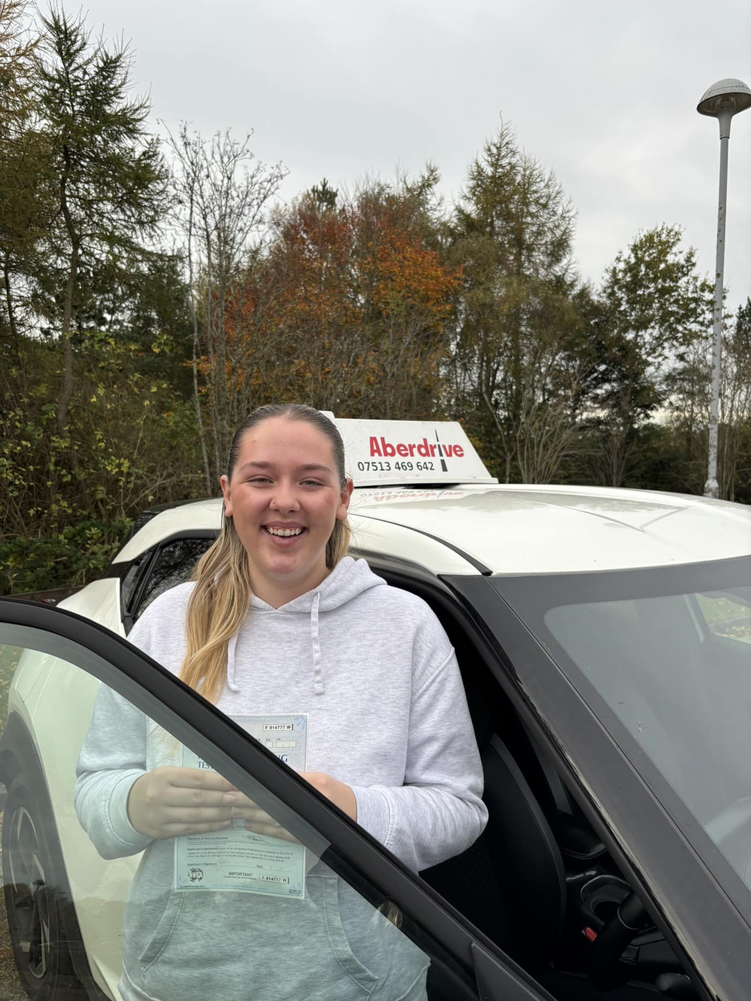 A young woman with blonde hair wearing a gray hoodie holding a driver's license, standing next to a white car with an Aberdrive driving school sign on top, outdoors with trees and an overcast sky in the background.