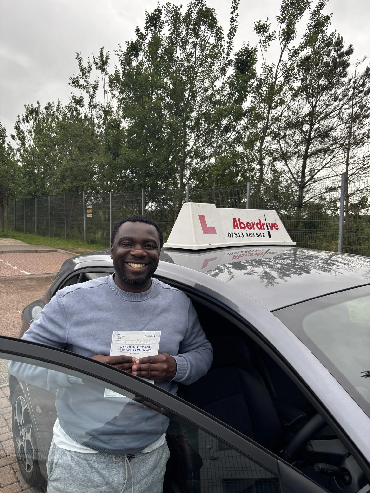 A man smiling and holding a practical driving test pass certificate standing next to a car with an 'Aberdrive' driving school sign on top, outdoors with trees and a fence in the background.