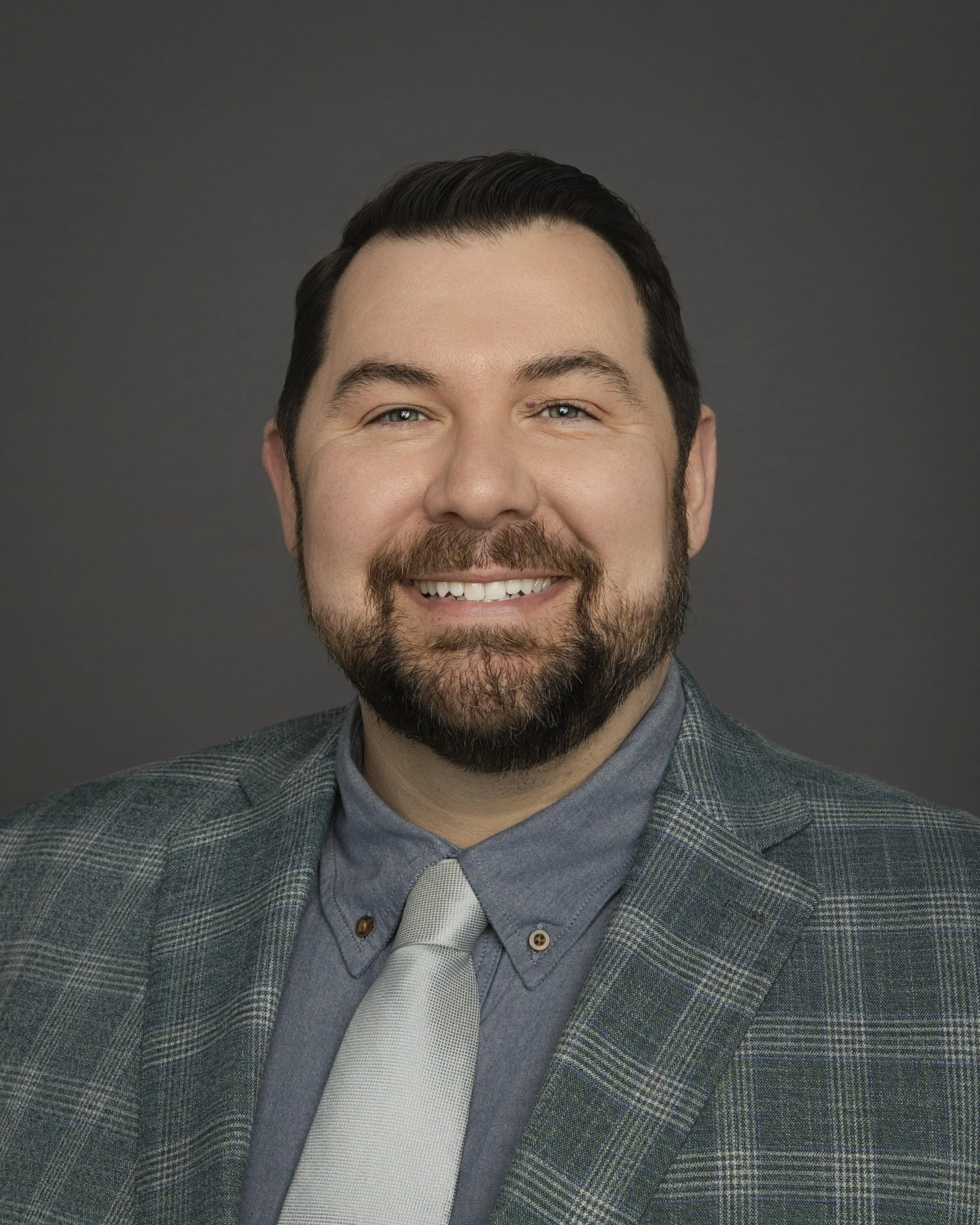 Headshot of a smiling man with dark hair, beard, wearing a plaid blazer, gray shirt, and light-colored tie against a dark backdrop.