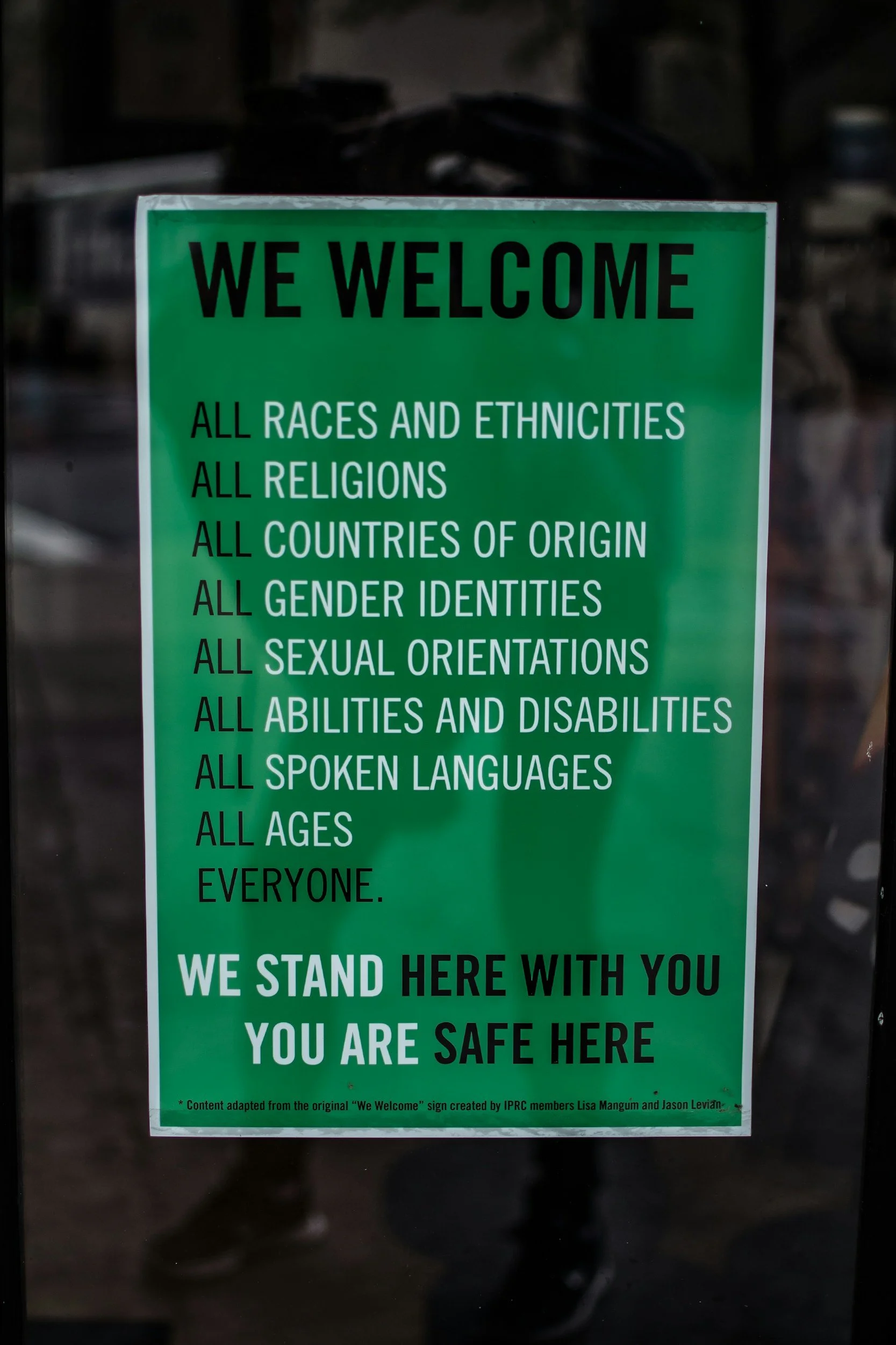 A green sign with black and white text welcoming everyone and promoting inclusivity across races, ethnicities, religions, nationalities, gender identities, sexual orientations, abilities, languages, and ages, indicating the place is safe and welcoming.