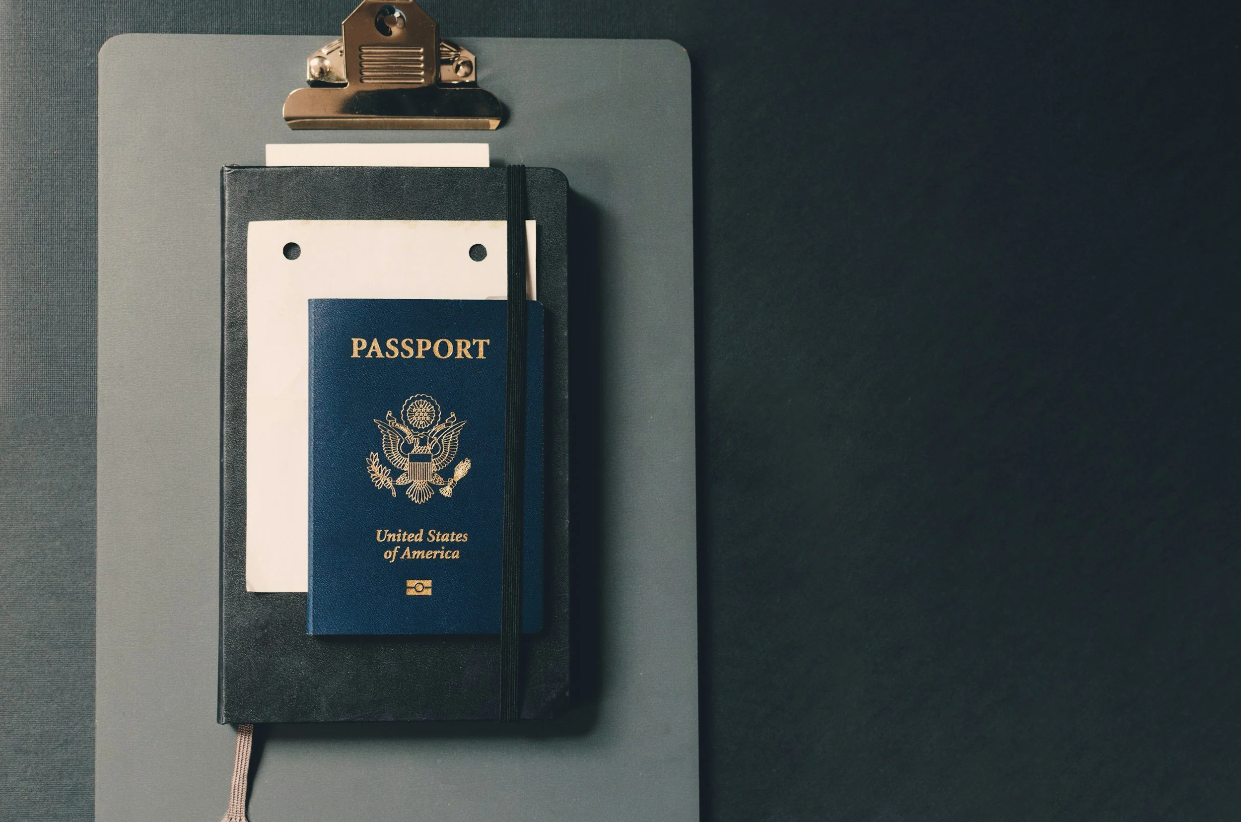 A United States passport attached to a black notebook with a white elastic band, on a gray clipboard, on a dark surface.