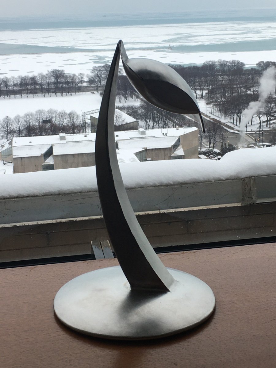A modern stainless steel abstract sculpture resembling a flower with a large petal and curved stem, placed on a windowsill overlooking a snowy landscape with buildings, trees, and a frozen body of water in the background.