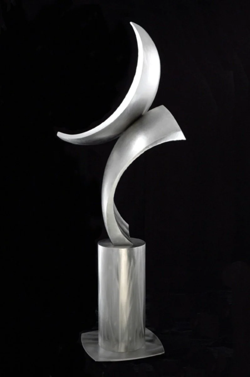 Sculpture of a stylized crescent moon and abstract elements on a cylindrical base, which rotates, set against a black background.