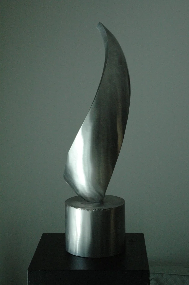 A modern, abstract sculpture made of stainless steel, with a curved, elongated shape, mounted on a cylindrical base.
