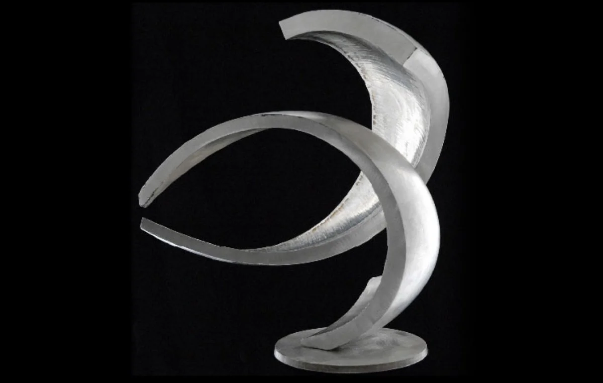 Abstract stainless steel sculpture, with compound curves, mounted on a circular base, set against a black background.