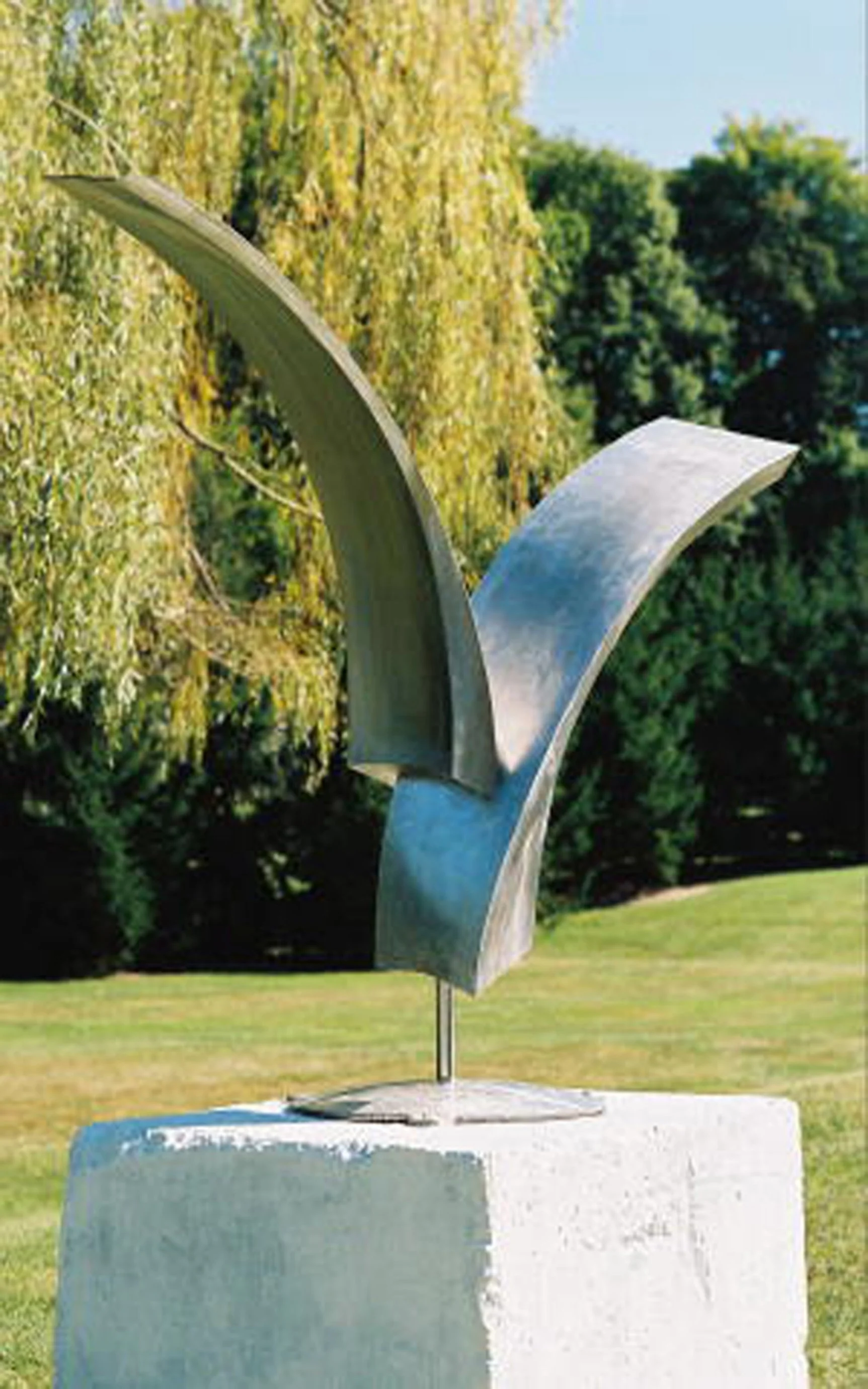 Abstract stainless steel sculpture outdoors surrounded by green trees and grass.