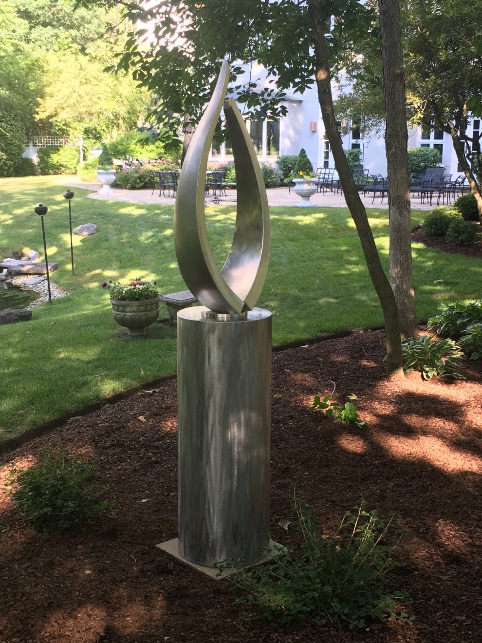 Modern abstract stainless steel sculpture in a landscaped garden with trees, plants, a flower bed, and a seating area in the background.