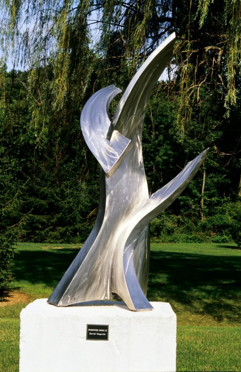 A modern outdoor sculpture made of stainless steel  with abstract, flowing shapes, mounted on a white pedestal in a park setting with trees and grass in the background.