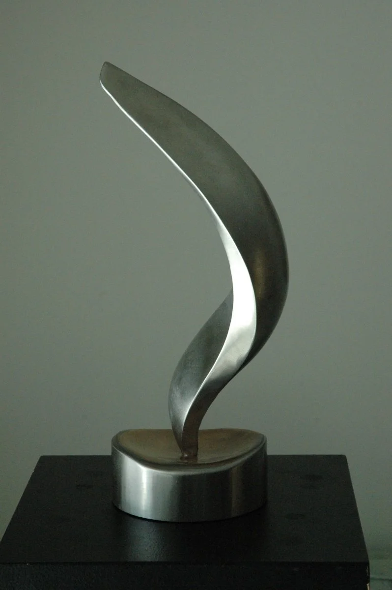 A modern abstract stainless steel sculpture with a twisted, curved design, placed on a dark surface against a plain background.