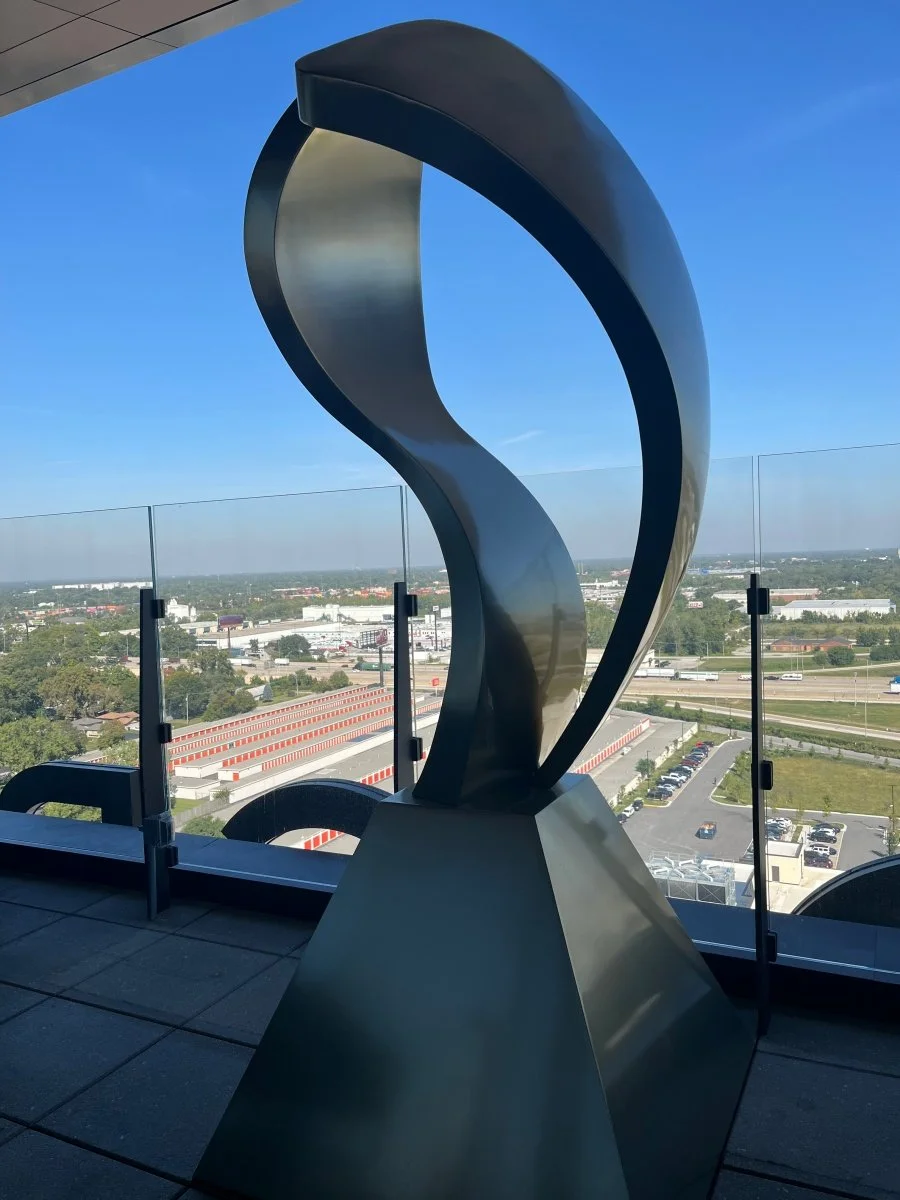 A large modern stainless steel sculpture on a balcony with a glass barrier, overlooking a cityscape with buildings, roads, and parking lots under a blue sky.