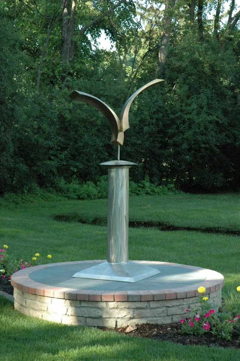 A modern abstract stainless steel sculpture, mounted on a tall cylindrical base, located on a brick and stone circular platform in a grassy garden with trees and flowers in the background.