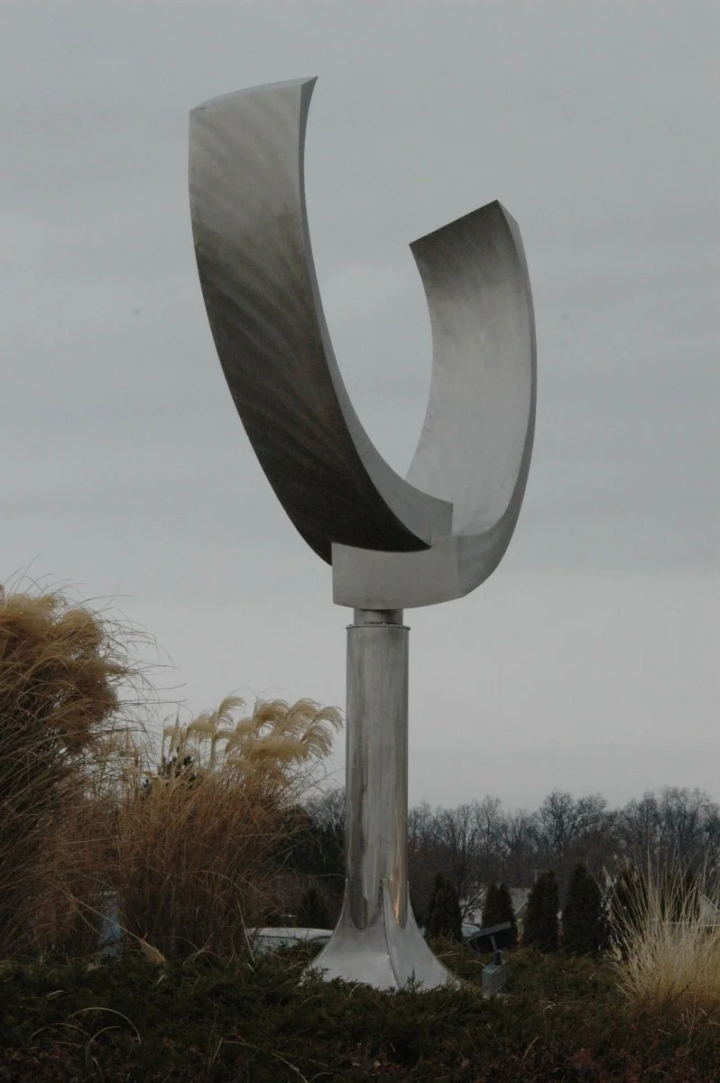 A large, modern stainless steel sculpture with curved shapes, set outdoors against a gray sky, surrounded by dry grasses and trees.