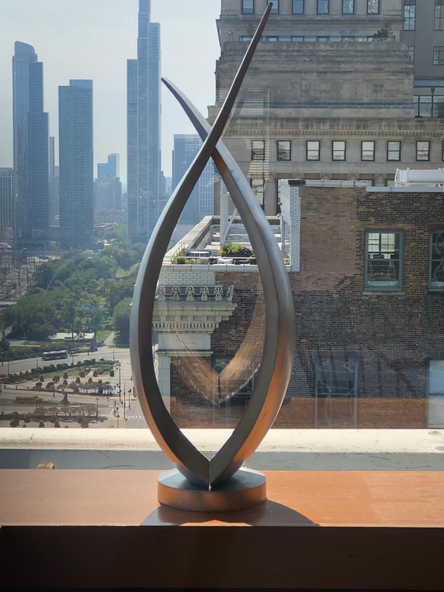 Abstract stainless steel sculpture with teardrop shape on a windowsill, cityscape with skyscrapers and a park in the background