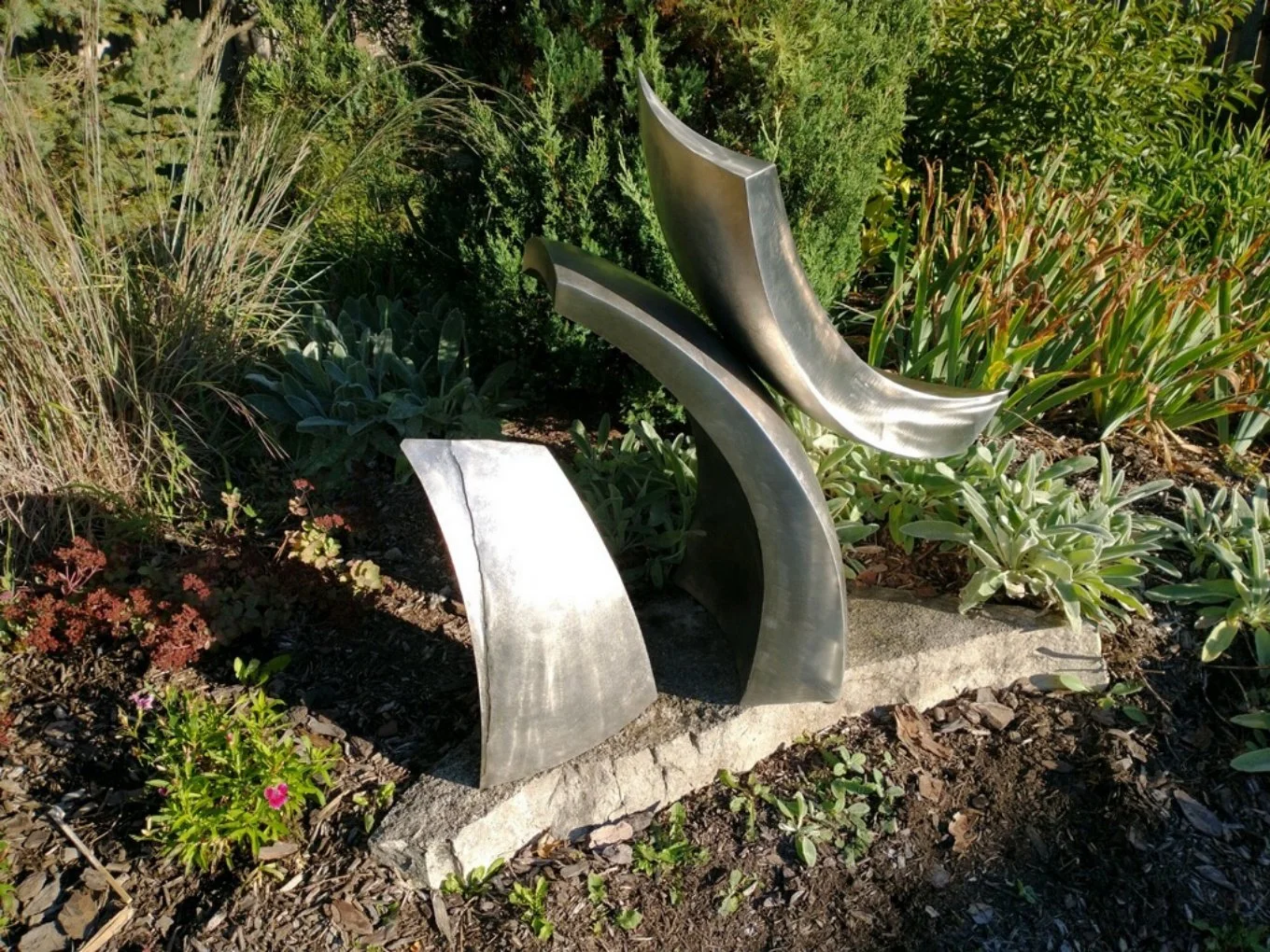A modern abstract stainless steel sculpture, placed on a stone plinth in a garden with various green plants and flowers.