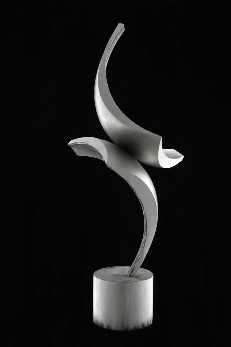 Modern abstract sculpture made of stainless steell with compound curves, flowing shapes on a cylindrical pedestal against a black background.