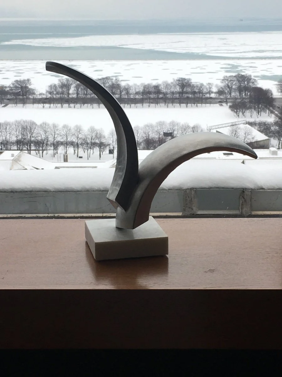 Abstract stainless steel sculpture of wings mounted on a marble base, displayed on a wooden table with snowy landscape and ice-covered water in the background