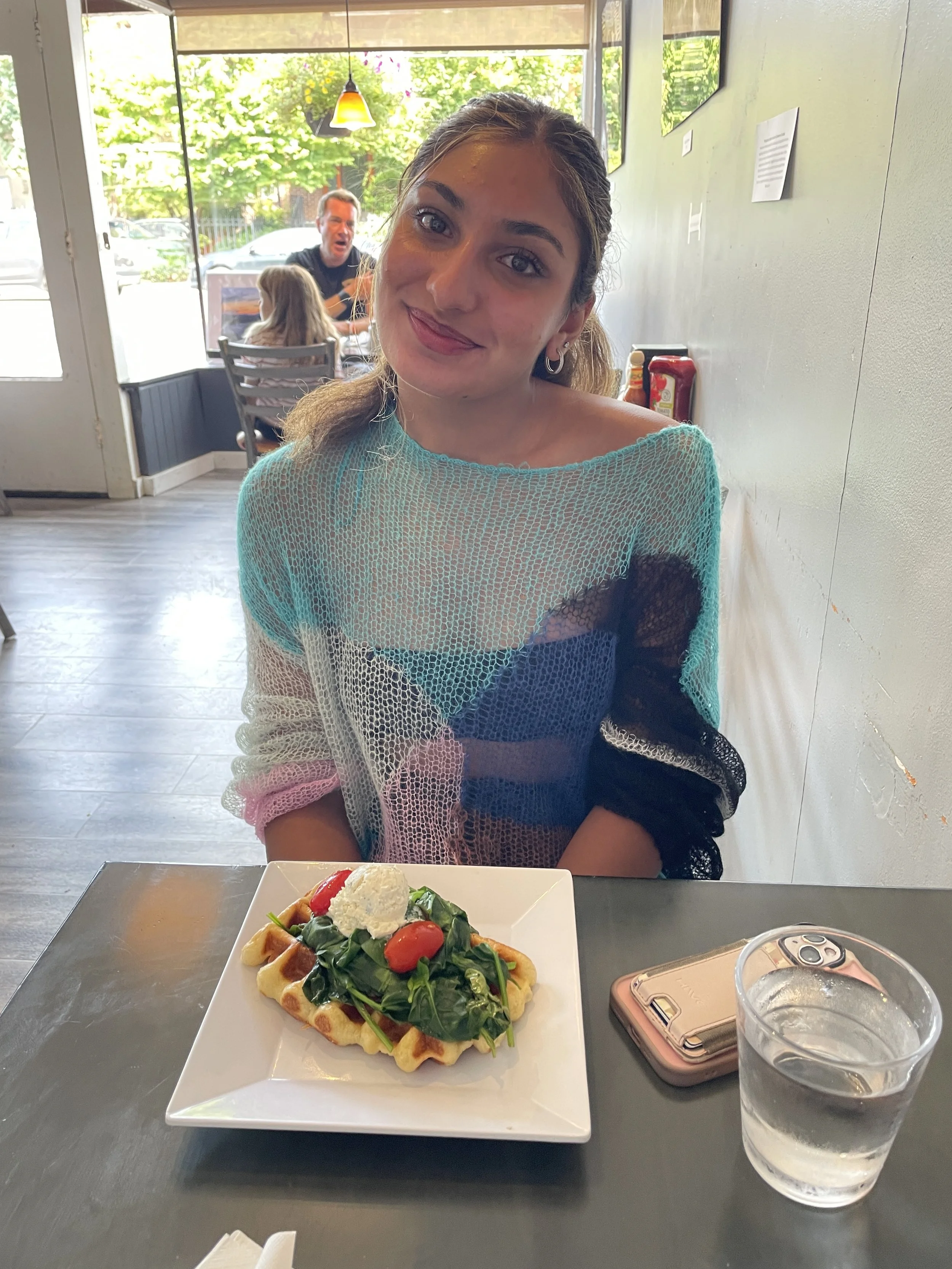 A young woman with light skin and brown hair smiling at the camera, sitting at a table with a plate of waffle topped with greens, tomatoes, and a dollop of cream, in a bright restaurant.