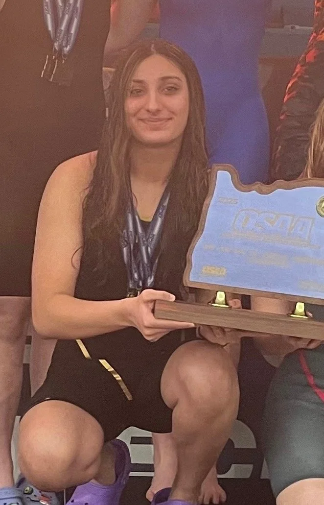 A young woman kneeling on one knee, holding a wooden plaque or award. She has long brown hair, is smiling, and is wearing a black athletic outfit with purple Crocs. She is in an indoor setting, possibly at a sporting event or award ceremony.