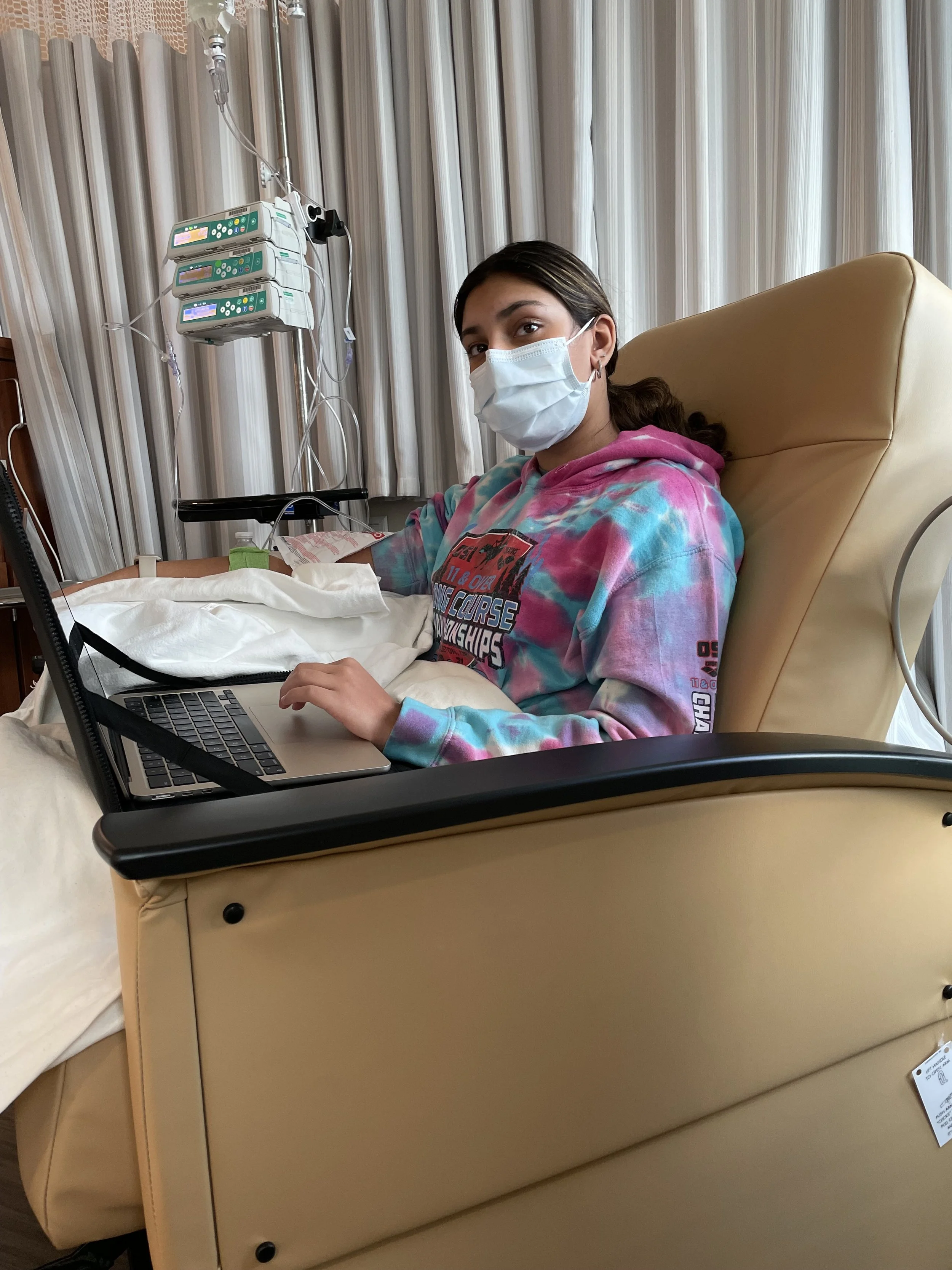 A young woman with brown hair wearing a pink and blue tie-dye hoodie and a face mask, sitting in a hospital bed with a laptop on her lap, connected to medical monitors.