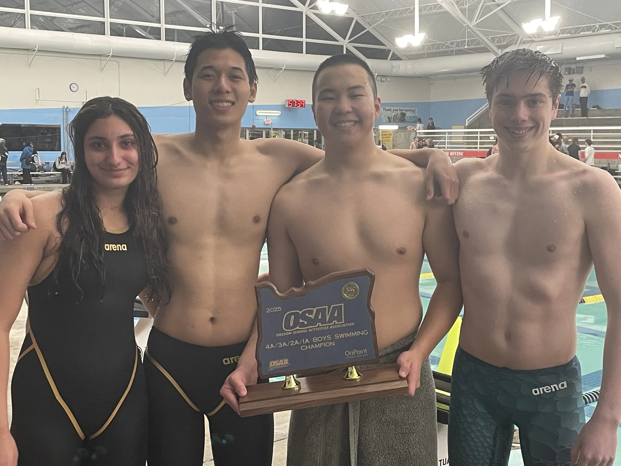 Four young swimmers standing together at an indoor swimming pool, holding a trophy for being boys swimming champions. They are smiling and wearing swimming trunks, with one woman in a black swimsuit on the left.