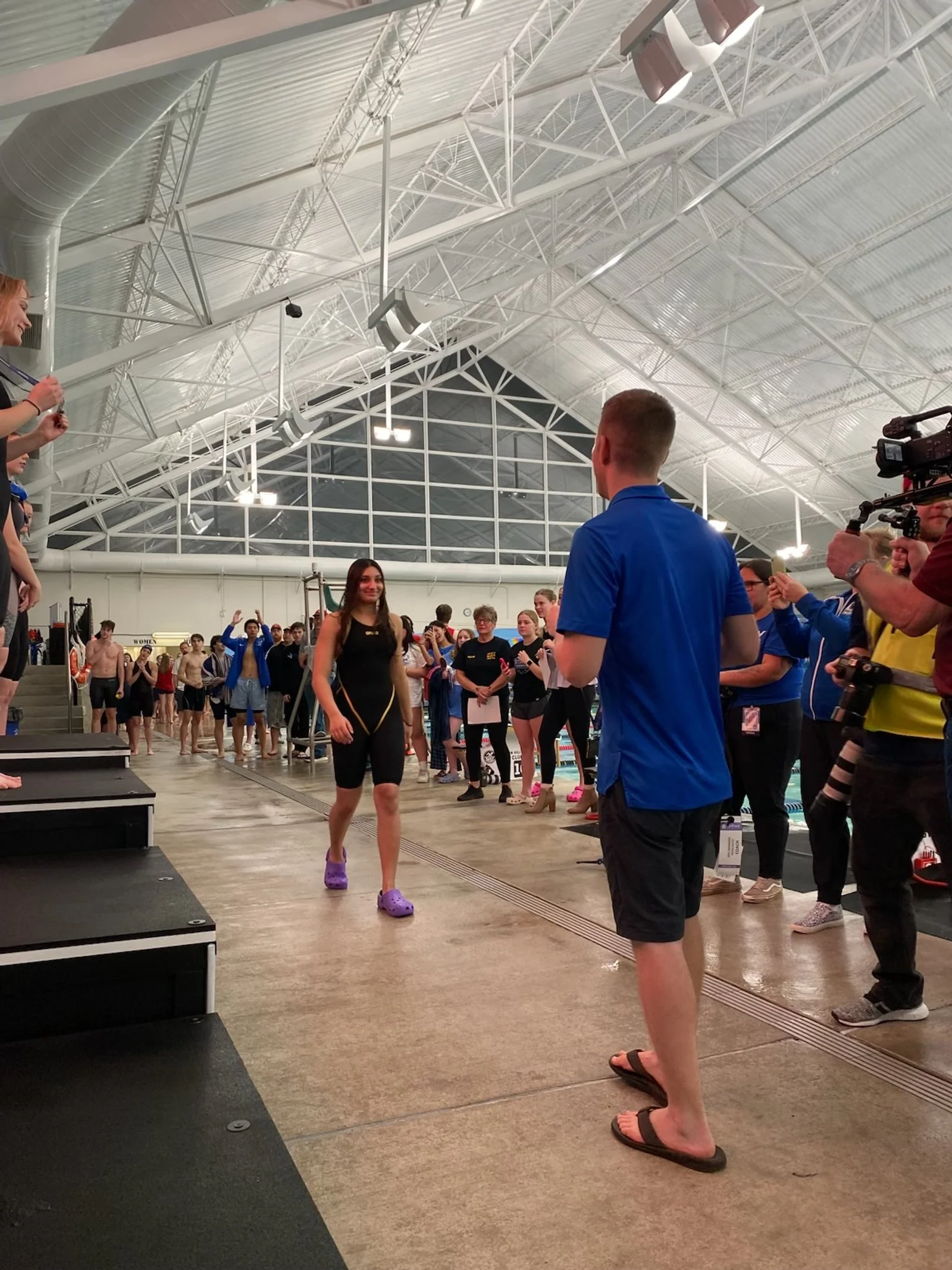 A young woman wearing black athletic wear and purple Crocs walking across a stage while smiling. She appears to be at a swimming pool or sports facility, with many people in the background watching and some taking photos. The high ceiling has a metal framework and large lights.