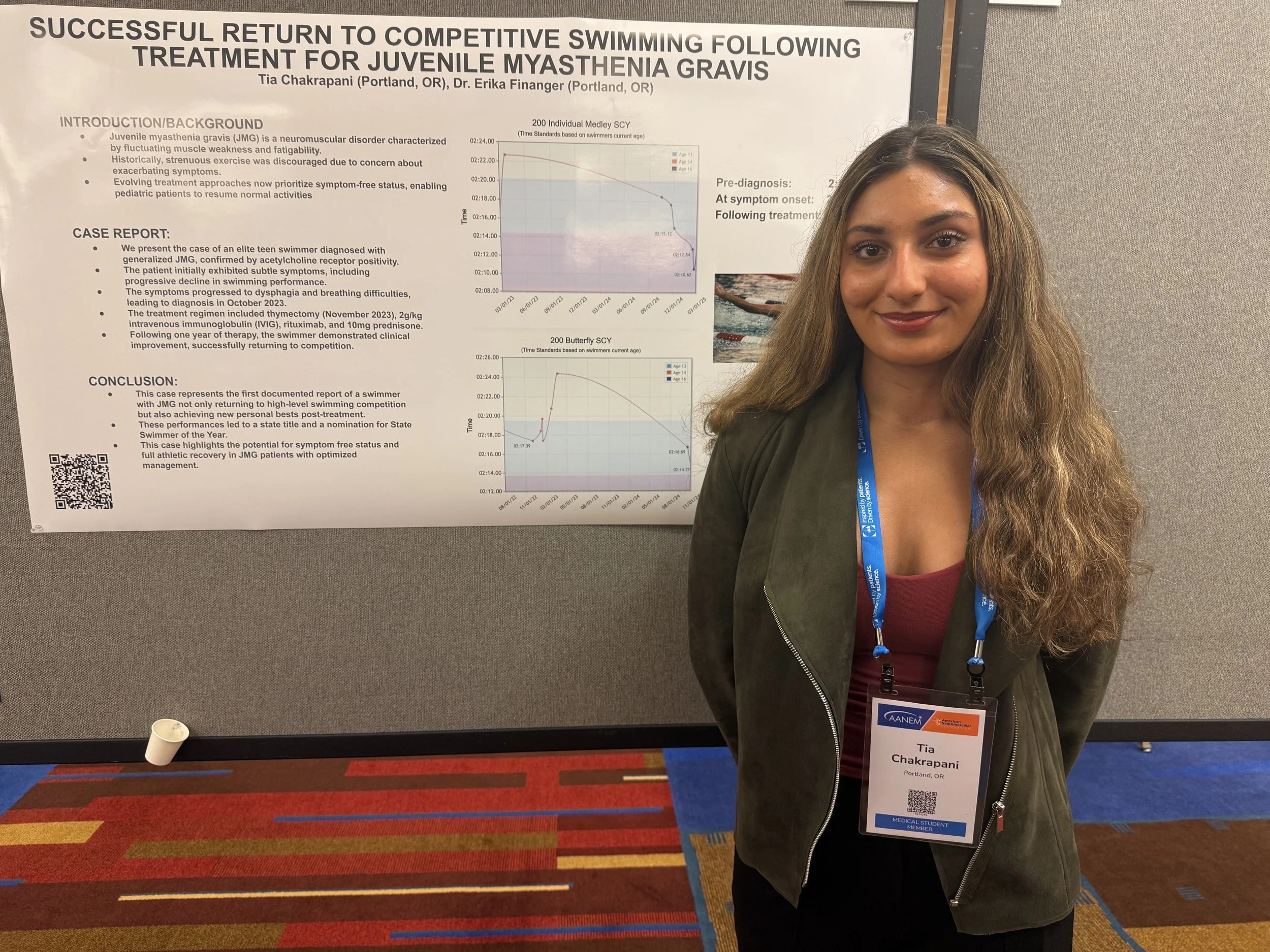 Young woman with long wavy hair standing in front of a research poster at a conference. She is wearing a name badge that says Tia Chakrapani from Portland, OR, and a dark green jacket over a maroon top. The poster behind her discusses a successful return to competitive swimming following treatment for juvenile myasthenia gravis.