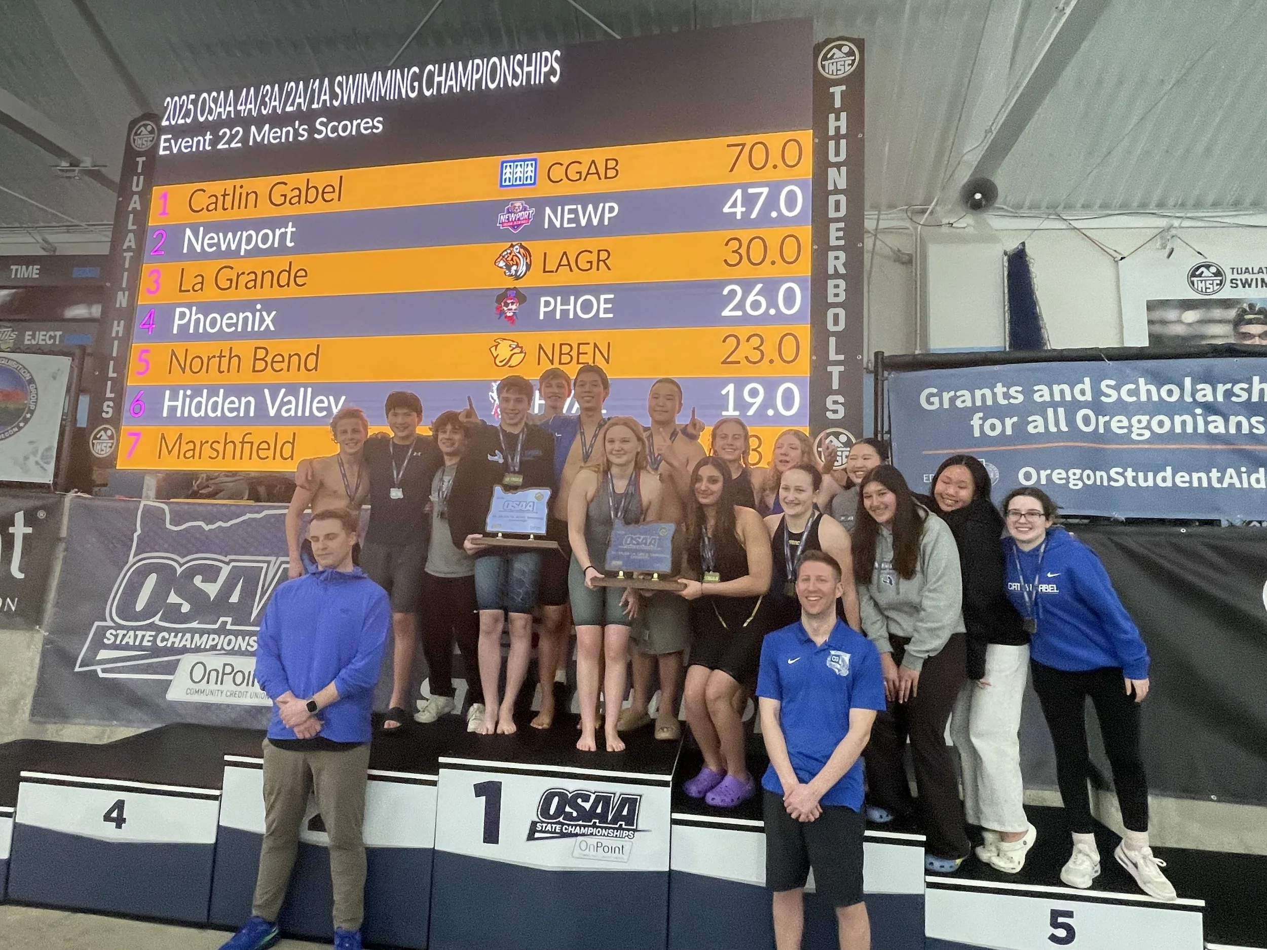 Swimmers and coaches celebrating on a winners' podium at an Oregon swimming championship, with a large scoreboard displaying the top teams and scores behind them.