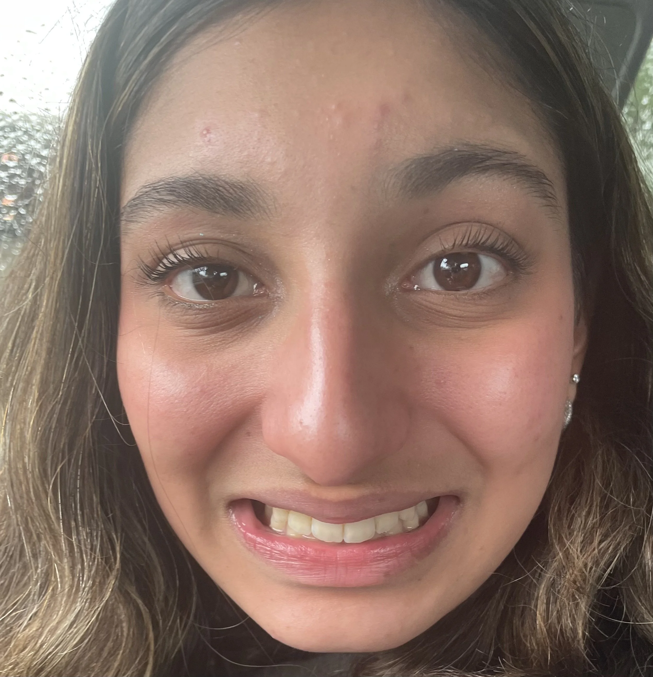 Close-up of a young woman's face smiling indoors, showing brown eyes, light brown hair, and small earrings.