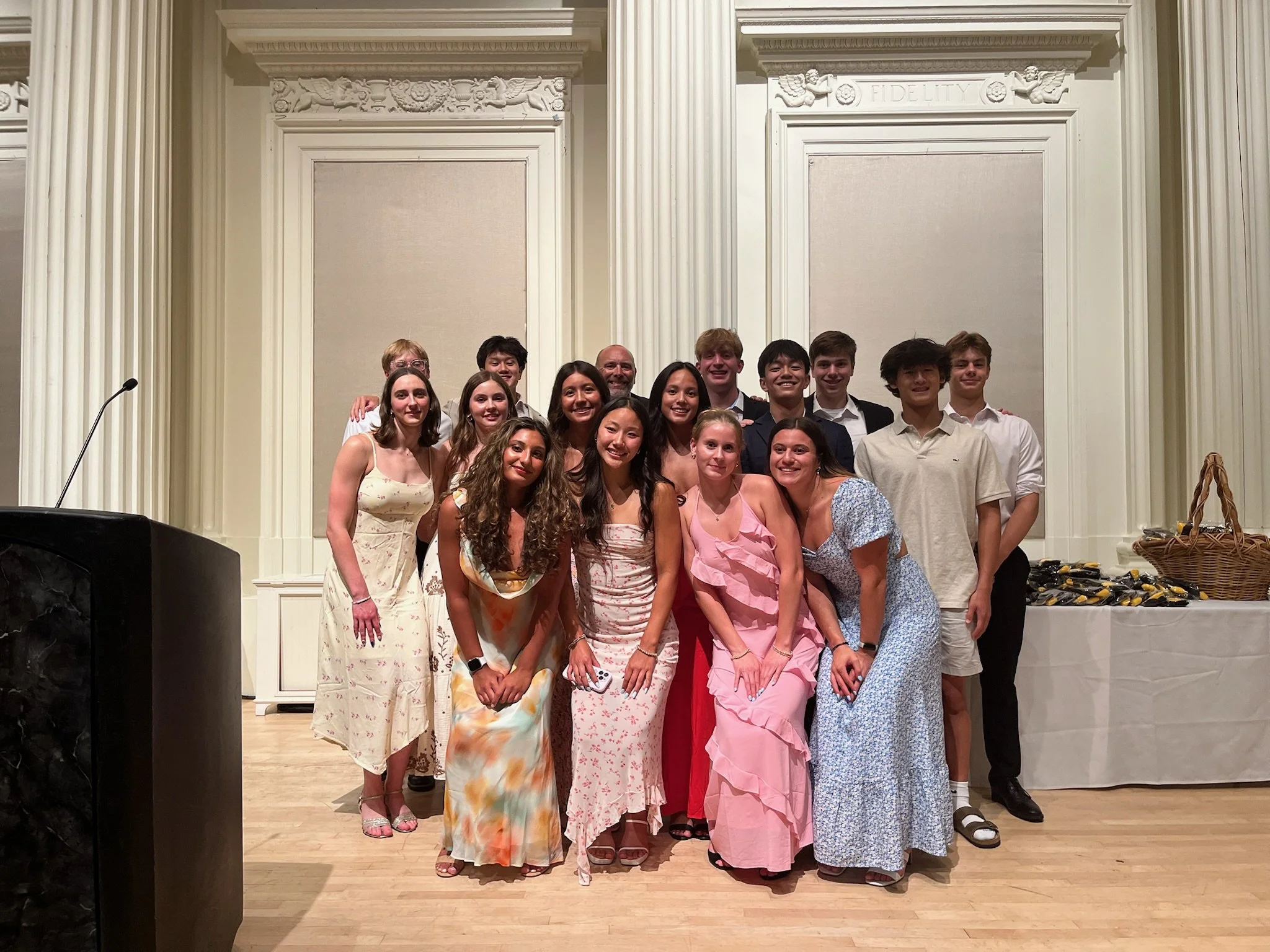 A group of young people dressed in formal and semi-formal attire posing on stage in front of a classical white architectural backdrop at a celebratory event.