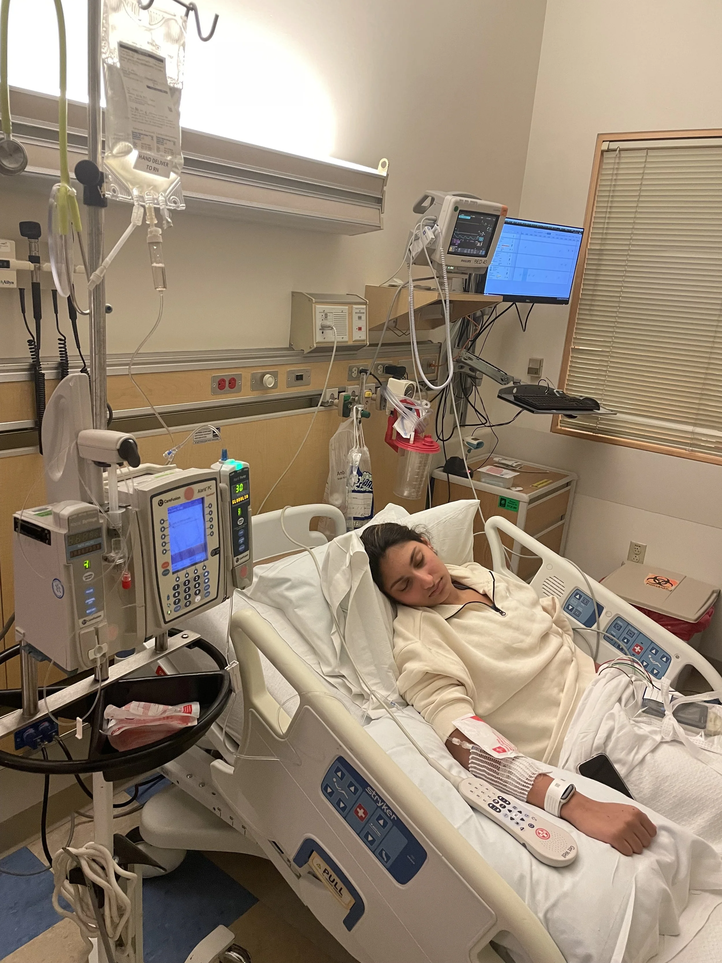 A woman lying in a hospital bed connected to medical equipment, including monitors and IVs, in a hospital room.