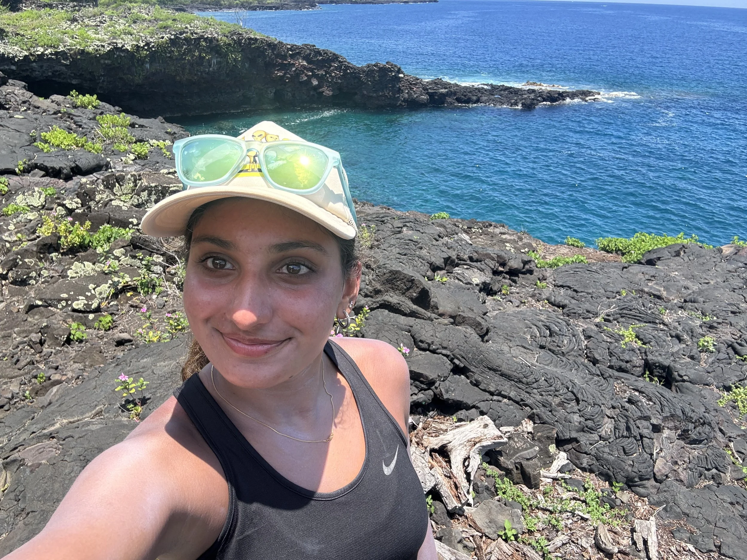 A smiling woman taking a selfie on a rocky coastline with vibrant blue ocean water in the background. She is wearing a black tank top, a light-colored cap with yellow sunglasses resting on top, and small earrings.
