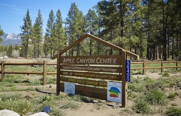 Sign for Apple Canyon Center surrounded by trees, with a wooden fence and mountains in the background.