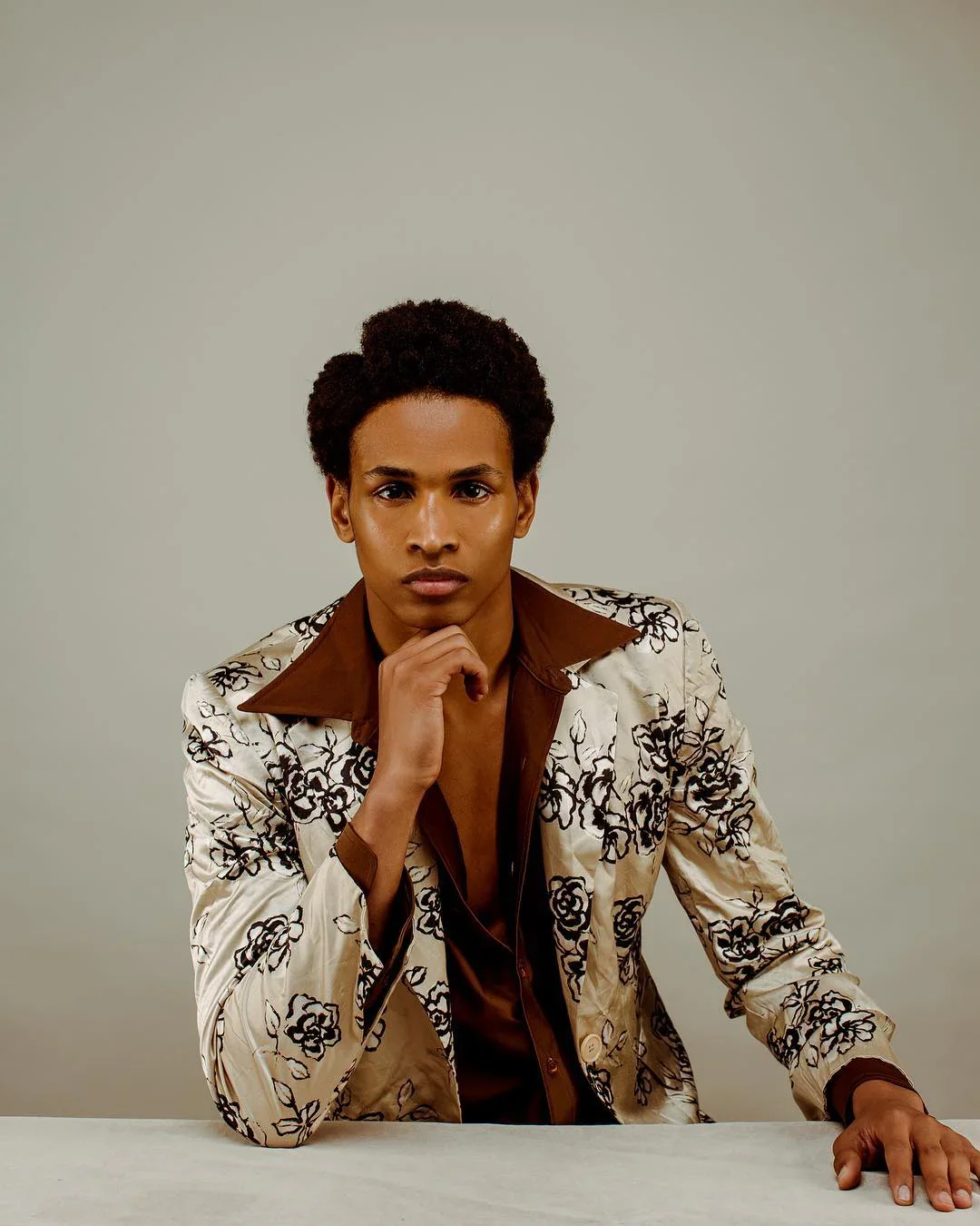 A young man with dark skin and short curly hair sitting at a table against a plain background, wearing a cream-colored jacket with black floral embroidery and a brown shirt. He is resting his chin on his left hand and looking directly at the camera.
