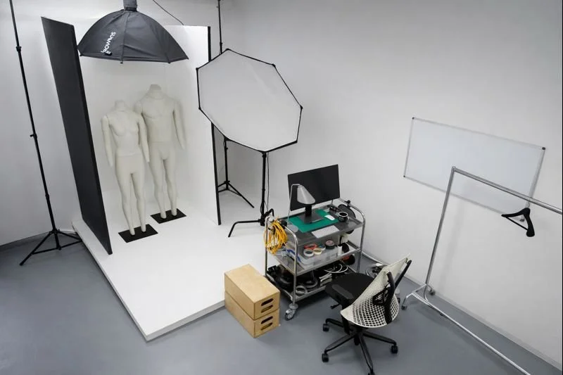 Photography studio setup with two mannequins on a white platform, background with umbrellas, softboxes, and studio lighting equipment, with a black chair, rolling cart with photography accessories, a dumbbell-shaped object, and a whiteboard in the room.