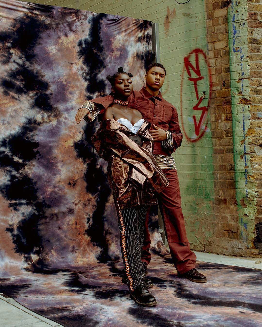 Two young people standing in front of a tie-dye backdrop and graffiti-covered brick wall, dressed in fashion-forward, edgy clothing.