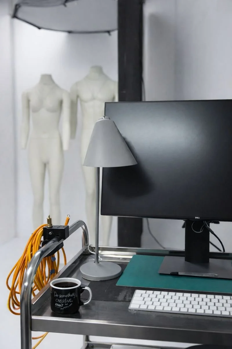 A workstation with a desktop computer monitor, a keyboard, a lamp, a coffee cup, usb c cable connection and mannequin torsos in the background.
